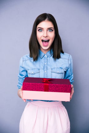 Surprised woman holding gift and looking on camera over gray background